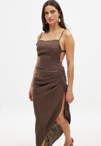 Ruskea pellavamekko, jossa on ohuet hihnat, kappaleenmyötäinen leikkaus, kippurainen hame, korkea halkio, teksturoitu kangas, yhdistettynä valkoisiin pisaramuotoisiin korvakoruihin.