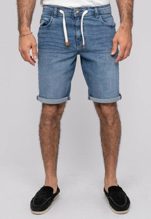 Homme portant un short en jean bleu retroussé avec un cordon blanc, une chemise blanche et des chaussures noires faciles à enfiler, debout devant un fond uni.