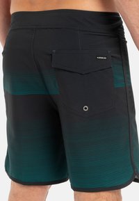 Sorte svømmeshorts med teal horisontale striper, elastisk midjebånd og en baklomme med klaff og øyelet-detalj.