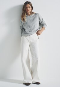 Maglione grigio chiaro con scollo tondo, abbinato a pantaloni bianchi svasati e scarpe marrone scuro con punte appuntite, su sfondo bianco semplice.