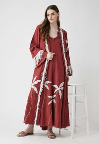 Robe longue de couleur rouille avec broderie de feuilles blanches, bords frangés et manches amples, portée avec des sandales à glisser marron.