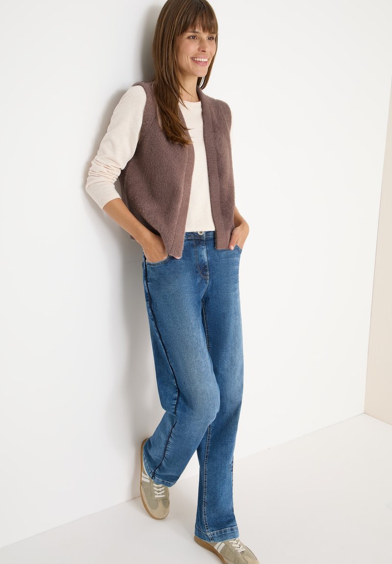 Frau lächelt, lehnt an einer Wand, trägt ein beiges Langarmshirt, eine braune, flauschige Weste, blaue Jeans und beige Sneaker mit weißen Streifen.