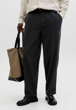 Person iført sorte bukser, sort bælte, beige skjorte, sorte sko, holder brun og sort shopper med den ene hånd i lommen.