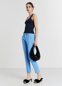Marineblaues Tanktop, blaue maßgeschneiderte Hose, schwarze Handtasche und schwarze Schuhe mit Blumenverzierung. Das Model steht vor einem neutralen Hintergrund.