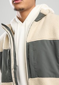 Fleecegefütterte Zip-Jacke mit einer Farbkombination aus Creme und Olivgrün, die weiche Texturen mit kontrastierenden Stoffpaneelen kombiniert.