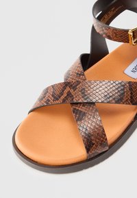 Sandalias de patrón de serpiente marrón con correas cruzadas, plantilla naranja suave y suela negra con un detalle de hebilla dorada.