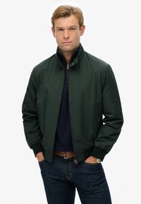 Donkergroen bomberjack met een ritssluiting aan de voorkant, drukknoopkraag en ribgebreide manchetten. Draag het over een marineblauw topje, gecombineerd met blauwe jeans. Geen zichtbare branding.