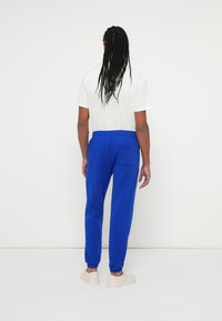 Ανδρικές μπλε φόρμες με ελαστική μέση, μανσέτες στους αστραγάλους και πίσω τσέπη; φορεμένες με λευκή μπλούζα και ανοιχτόχρωμα παπούτσια.