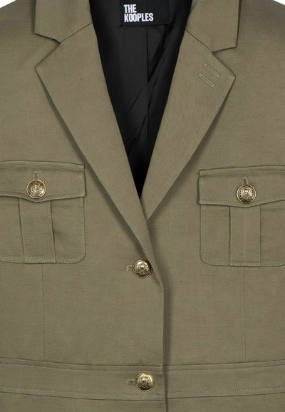 Olivgrüner Blazer aus strukturiertem Stoff mit zwei aufgesetzten Taschen vorne, goldenen Knöpfen und einem Reverskragen-Design.
