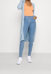 Cárdigan azul claro con frente abierto, combinado con una camiseta sin mangas naranja y jeans ajustados de lavado claro. Zapatillas blancas completan el conjunto.