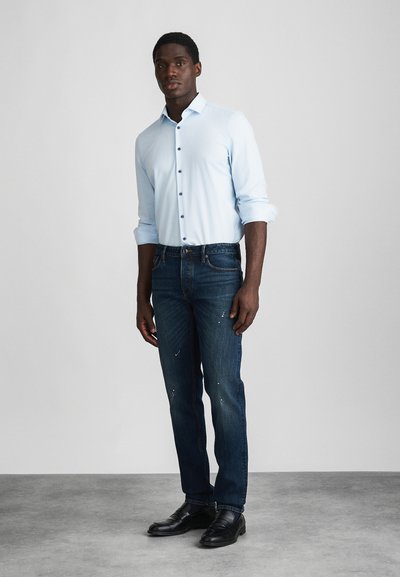 Mann står iført lys blå skjorte med knapper og oppbrettede ermer, mørkeblå jeans med malingssprut, og svarte finssko på grått gulv.