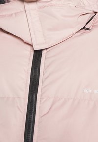 Ljusrosa puffjacka med hög krage, utrustad med en svart dragkedja framtill och en logotyp i vitt på nedre högra sidan.