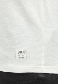 White cotton t-shirt with a textured finish and a rectangular fabric label on the lower left, displaying "!SOLID DENMARK EST.1999".