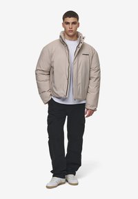 Jachetă puffer bej cu guler înalt, fermoar frontal și logo brodat. Purtați peste o cămașă albă, asortată cu pantaloni cargo negri și teniși albi.