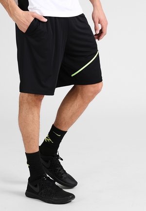 Mężczyzna noszący czarne sportowe spodenki z neonowym paskiem, czarne skarpetki i czarne buty sportowe, stojący z jedną ręką w kieszeni.