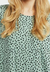 Mintgrön topp med lös passform, rund halsringning och ett svart prickigt mönster över hela tyget. Slät textur med en avslappnad design.