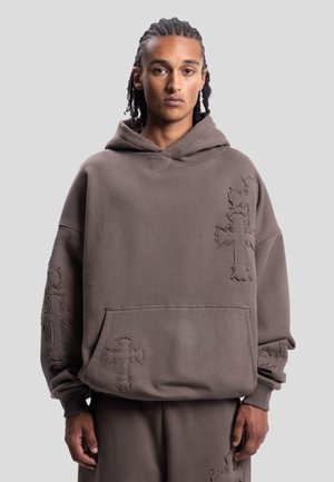 Giovane uomo con capelli intrecciati che indossa una felpa marrone larga e pantaloni con motivi a croce cuciti sulle maniche e sul davanti.