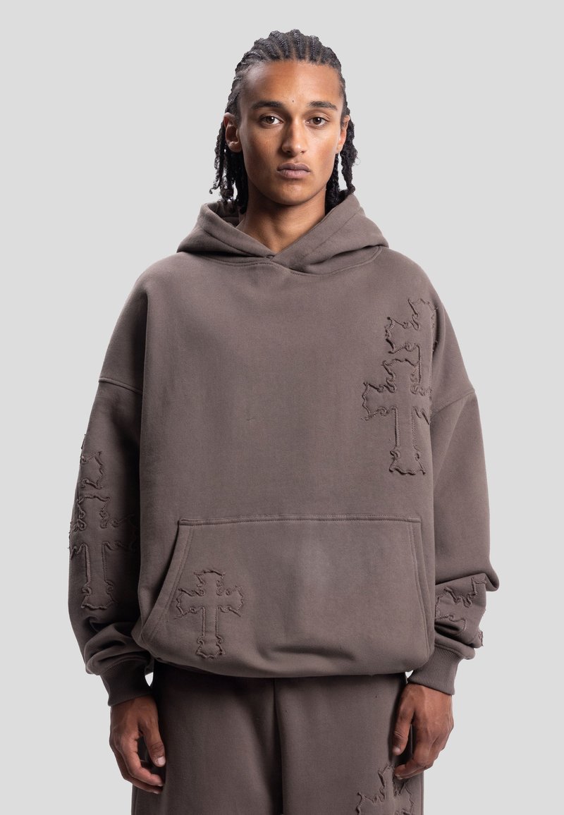 Jeune homme aux cheveux tressés portant un sweat à capuche marron ample et un pantalon avec des motifs de croix cousus sur les manches et le devant.