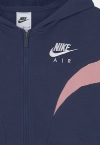 Námořnický modrý zipový svetr s růžovým zakřiveným akcentem a bílým vyšitým logem "Nike Air" na levé hrudi. Měkký bavlněný materiál.