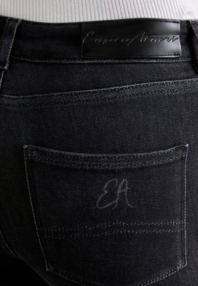 Mustat farkut, joissa on sileä pinta, taka-tasku ja kohokuvioitu "Emporio Armani" -merkki vyötäröllä. Huomaamaton "EA" kirjontayksityiskohta.