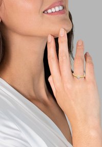 Anello d'oro con un singolo gemma trasparente, indossato sulla mano destra. Fascetta di metallo liscia, posizionata su uno sfondo grigio chiaro.