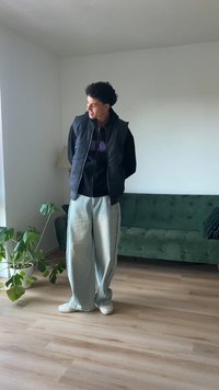 Svart puffervest over svart hettegenser, lyseblå vide bukser og hvite sneakers. Innendørs setting med grønn sofa og stueplante.