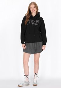 Fekete kapucnis pulóver csillogó grafikával, hosszú ujjú, elülső zsebbel, szürke pliszírozott szoknyával és ezüst tornacipővel, fűzős zoknival párosítva.