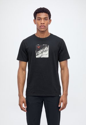 Un homme pose avec un T-shirt noir orné d’un petit motif graphique, devant un fond de studio blanc uni.
