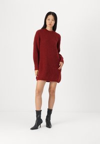 Knit suknelė iš gilaus raudono mišinio su tekstūruotu, raštuotu dizainu. turi ilgas rankoves ir aukštą apykaklę. Dėvima su juodais kulkšninius batais.