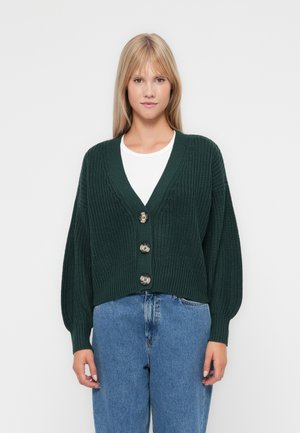Tmavě zelený pletený kardigan s výstřihem do V, zapínáním na knoflíky a oversize rukávy. V kombinaci se světle modrými denimovými kalhotami.