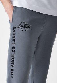 Calças de fato cinzas com um logótipo e texto bordados dos "Los Angeles Lakers" na lateral, apresentando uma textura de tecido suave.