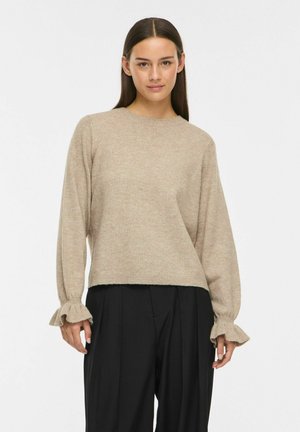Femme aux cheveux lisses et foncés porte un pull beige à manches longues avec des poignets à volants et un pantalon noir ample, posant devant un fond blanc.