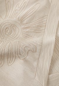 Tissu beige avec broderie de motif de soleil en fil plus clair. Texture lisse avec un éclat subtil, comportant des lignes courbes et des détails en spirale.