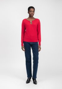 Rode gebreide trui met een sleutelgatsnit, voorzien van een metallic accent. Gecombineerd met donkerblauwe denim jeans en zwarte enkellaarsjes.