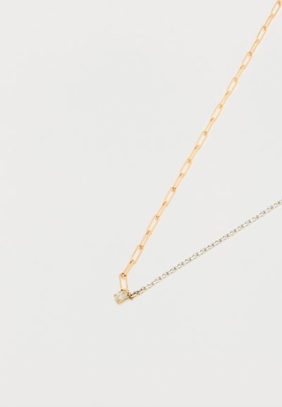 Δύο λεπτές αλυσίδες, μία χρυσή με επιμηκυσμένους συνδέσμους και μία ασημένια με μικρούς κυκλικούς συνδέσμους, συνδεδεμένες με ένα μικρό διάφανο πολύτιμο λίθο.