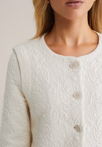 White quilted jacket with a textured paisley pattern, round neckline, and two buttons on the front. Soft fabric with a structured silhouette.