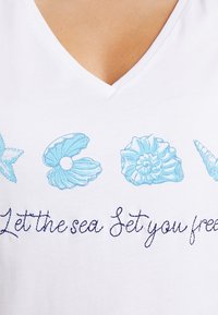 Bílý bavlněný tričko s výstřihem do V, zdobené modře vyšitými motivy mořských lastur a textem "Nech moře osvobodit tě" v modré nitě.