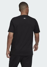 Muž zobrazený zezadu, který má na sobě černé tričko Adidas s logem na horní části zad a černé kalhoty na jednobarevném pozadí.