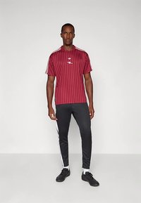 Maglietta a maniche corte con righe color granata e dettagli bianchi, abbinata a pantaloni neri a cono con righe laterali bianche. Scarpe da ginnastica nere.