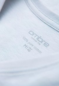 En lyseblå bomulds skjorte med et mærkat, der viser "ombre" branding. Mærket angiver "100% ren bomuld" og størrelsesdetaljer med grå skrift. En ensartet tekstur uden mønstre.
