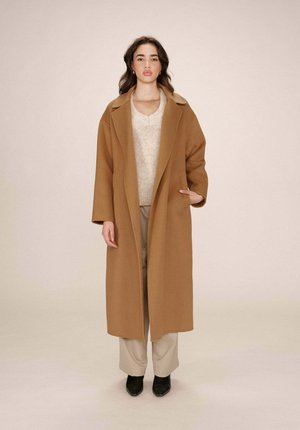 Long manteau en laine marron avec de larges revers, porté sur un pull beige et un pantalon clair, associé à des chaussures noires à bouts pointus.