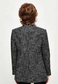 Černobílý tvídový blejzr s texturovaným povrchem, zdobený volánkovým límcem a třepeným lemem, střih na míru a dlouhé rukávy.