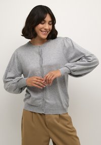 Gilet en tricot gris avec un col rond, une fermeture à boutons et des manches bouffantes. Le tissu a une texture douce, s'harmonisant avec un pantalon beige.