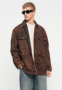 Brown distressed denim jacket with a contrasting black inner lining, two front pockets, and metal button details. Worn over a light shirt.