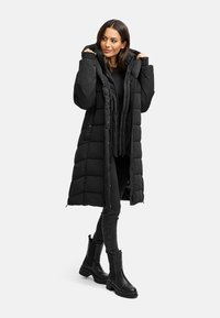 Schwarz gepolsterter langer Mantel mit Kapuze, vorderen Knöpfen und Taschen, kombiniert mit schwarzen eng anliegenden Hosen und Stiefeletten. Glatte Textur.