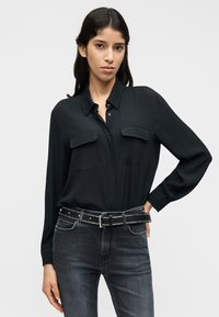 Μαύρο πουκάμισο με μακριά μανίκια που διαθέτει δύο τσέπες στο στήθος, γιακά με κουμπιά και ομαλή υφή. Συνδυάζεται με σκούρο τζιν και ζώνη με τρουκς.