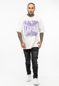 Hvid kortærmet t-shirt med et lilla grafisk design med ordet "TAPOUT." Modellen bærer slidte sorte jeans og hvide sneakers.