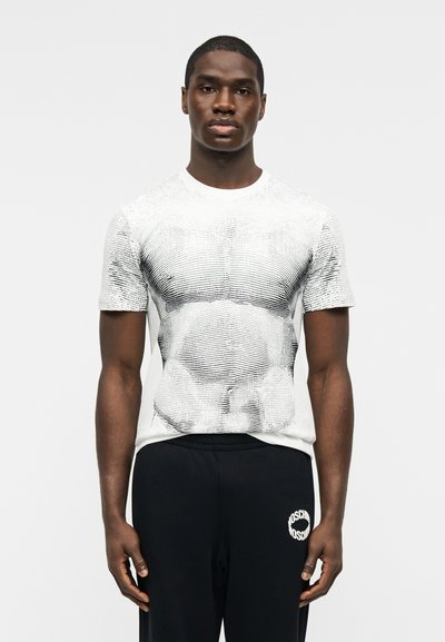 Άνδρας που στέκεται και κοιτάζει μπροστά, φοράει λευκό μπλουζάκι με μαύρες αφηρημένες γραμμές και μαύρο παντελόνι με λευκό κυκλικό λογότυπο.