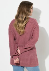 Przewymiarowany, długi sweter w kolorze pudrowego różu, z prążkowaną teksturą i bocznymi rozcięciami, zestawiony z jasnoniebieskimi dżinsami.