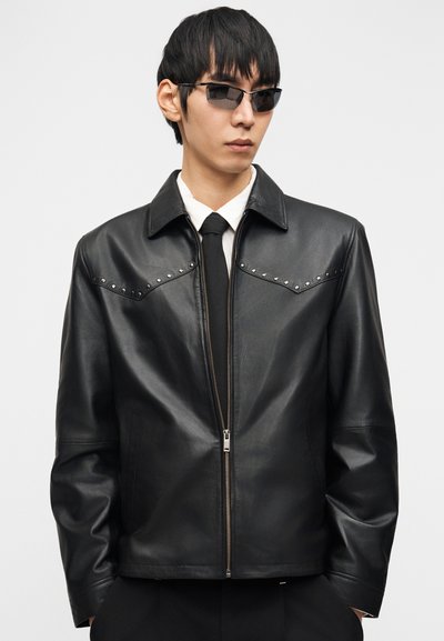 Άνδρας που φοράει μαύρο δερμάτινο μπουφάν με στρας, άσπρο πουκάμισο, μαύρη γραβάτα και στενά μαύρα γυαλιά ηλίου, μπροστά σε απλό λευκό φόντο.